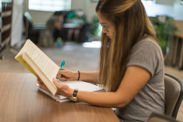 Student has a book open in her left hand and is writing on a notebook with her right.