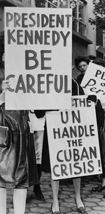 Historic black and white photo of group of women strikers for peace, holding signs that read "PRESIDENT KENNEDY BE CAREFUL" and "PEACE OR PERISH"