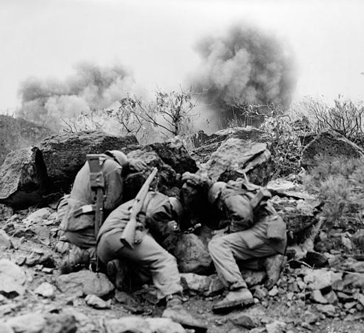 Historic black and white photo of Korean War soldiers covering up behind rocks to shield themselves from exploding mortar shells