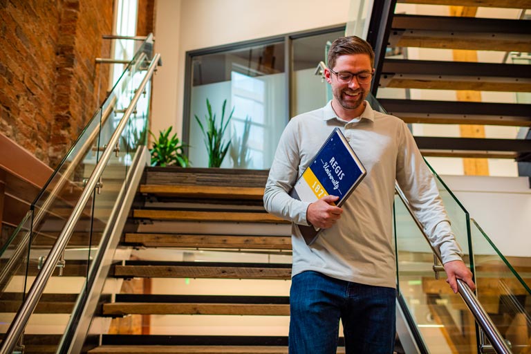 Adult student walks down stairs on the right hand side while smiling. Caring notebook that says Regis University 1877