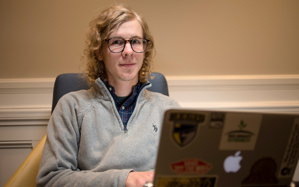 A student with glasses and curly hair sits with a laptop covered in stickers.