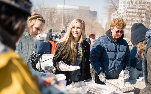 Regis University students volunteer at an outdoor food distribution event, serving meals to people on a cold day in an urban setting