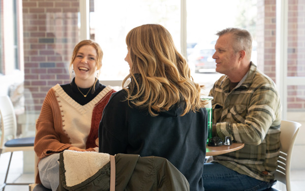 A Regis student with her parents sit at a table in a bright cafe, engaged in conversation while smiling.