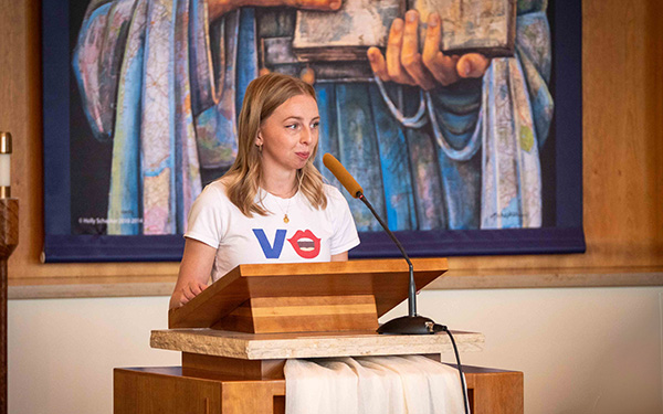 A young woman standing at a podium, speaking into a microphone. She is wearing a white shirt with the word "VOTE".