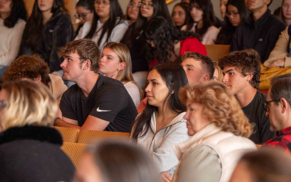 An engaged audience sitting closely together, attentively watching or listening to an event or presentation. The group includes people of various ages, seated in rows. The expressions of the attendees suggest focus and interest. The background shows additional individuals.