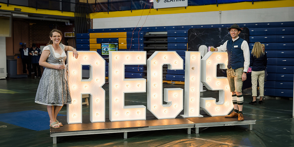 two attendees of Iggy Beer Fest wear traditional German clothing while holding tasting glasses and posing with a lighted Regis sign during blue and gold weekend in the field house