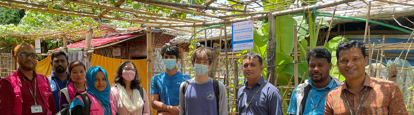 a diverse group of 10 care providers wearing a variety of backpacks, vest and lanyards stand among bamboo structures and jungle foliage in Myanmar