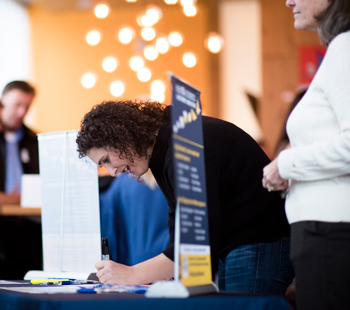 Person in the middle of frame writes on table. One person in the background on the left. One person in the right foreground