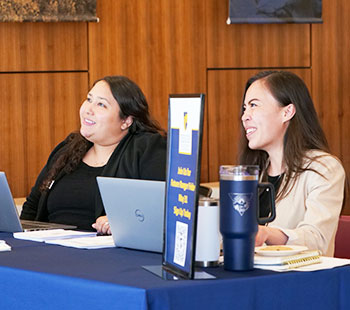Two admissions staff members sit at a table ready to answer questions at an event in the atrium of Clarke Hall on the Northwest Denver campus