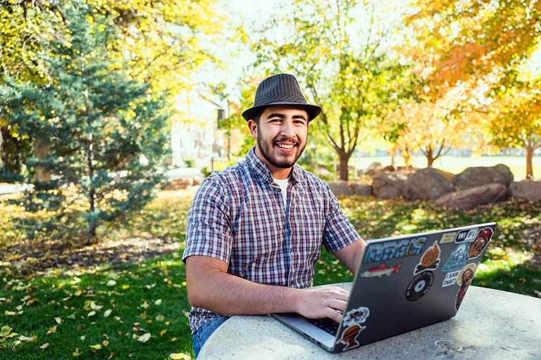 an adult student sits at an outdoor table working on his laptop