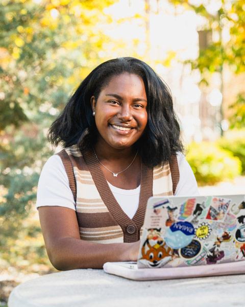 student smiling and sitting at an outdoor table on campus, on her laptop