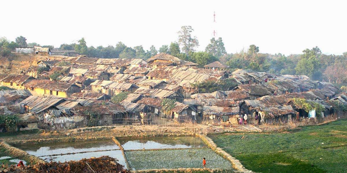 a camp of low structures built from a variety of materials and thatched roofs nestle together near a flooded field in Myanmar