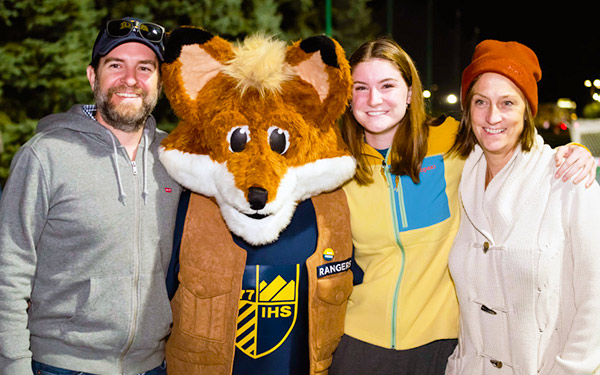 A Regis student and their parents pose for a photo with Regi the Fox during a night game on the match pitch