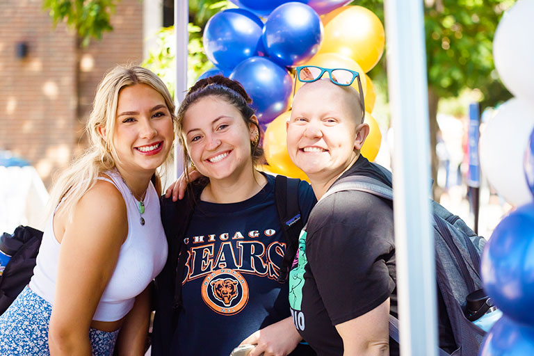 three Regis students pose for a photo while standing under a tent on the quad at blue and gold weekend on the Regis campus