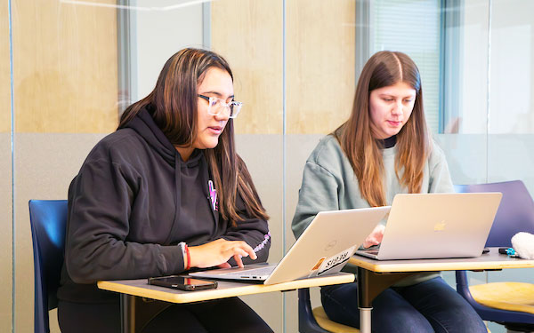 two students sit at desks and work on their laptops in a classroom in the Anderson College of Business and Computing