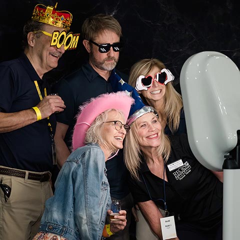 five attendees use props like silly glasses and whimsical hats while posing for a group photo at the photo booth