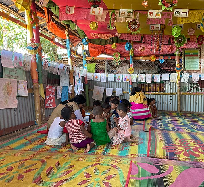 two teachers crouch on the floor with two groups of young children inside an open air bamboo structure draped with colorful fabric, banners and children's artwork in Myanmar