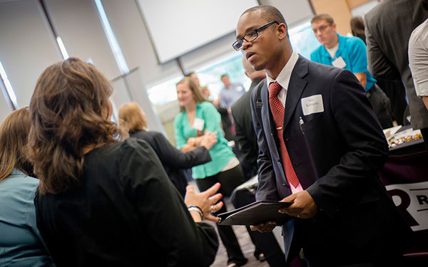 a young man wearing a suit talks to a potential employer in the crowded Mountain View Room during a career fair on the Regis campus