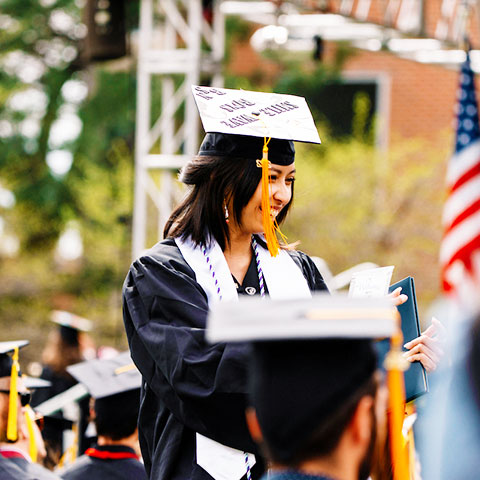 a graduate wearing regalia smiles as she walks to her seat holding her diploma cover at commencement ceremonies on the Quad on the Northwest Denver campus