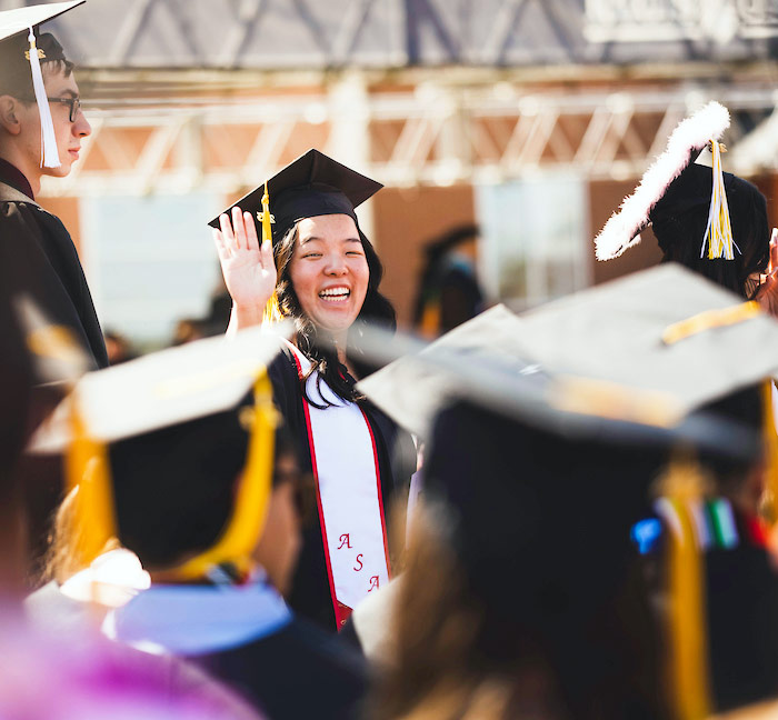 a student in commencement robes waves from among the crowd of graduates at commencement ceremonies on the regis campus