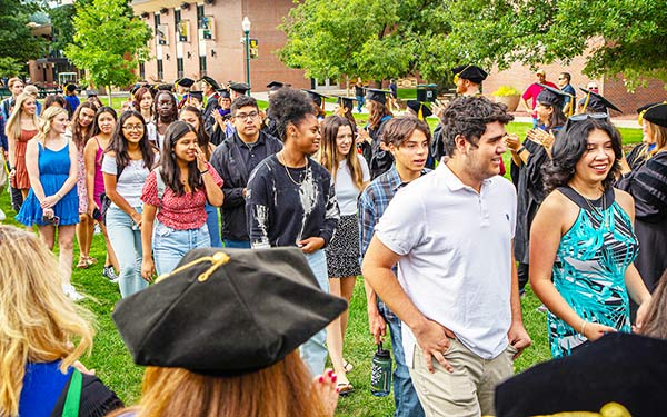 a marching band in jeans and t-shirts plays tuba, trumpets, saxophones, and base drums marches across the Quad while faculty in regalia cheer on the sidelines