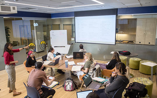 a small group of students sits in the innovation lab classroom in Clarke Hall as their professor talks and gestures to a powerpoint presentation
