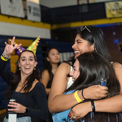 two attendees embrace while a third gives a peace sign while wearing two party hats in the fieldhouse during iggy beerfest