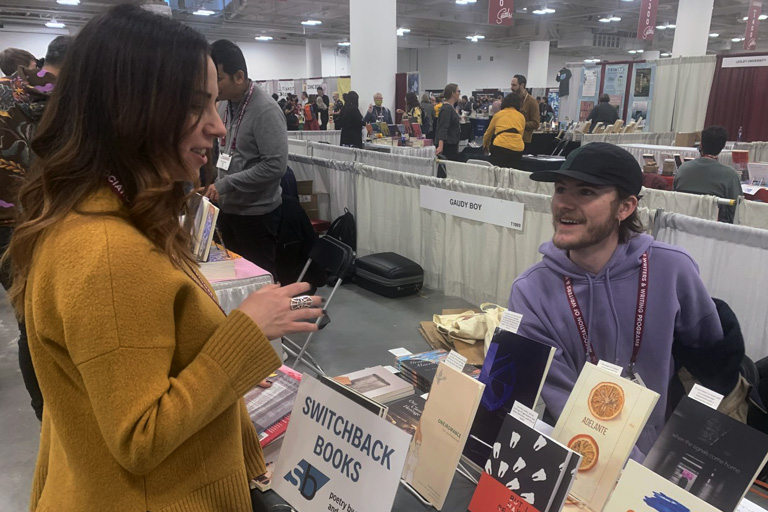 student behind booth titled "Switchback Books" with tables full of books on display, student is talking to a person who has approached the booth