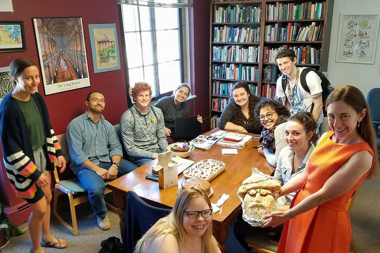 group of students and english department faculty members seated and standing around a table with food and other reading materials, all smiling at the camera