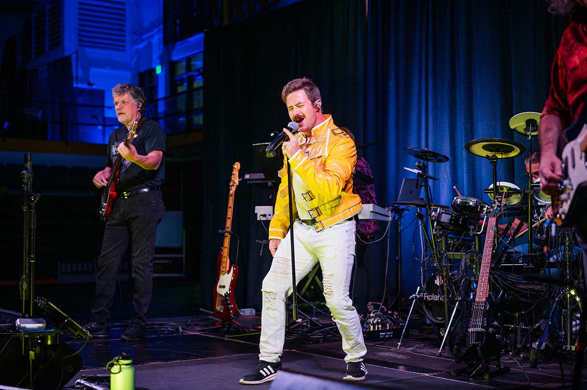 the lead singer of the fat bottom boys wears a Freddie-Mercury-esque yellow leather jacket while singing into a microphone on the stage in the fieldhouse. Guitarists flank him with the drummer behind and varios instruments, speakers and cables clutter the stage. 