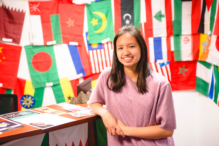 female student smiling and posed in front of desk, with a wall covered by international flags behind