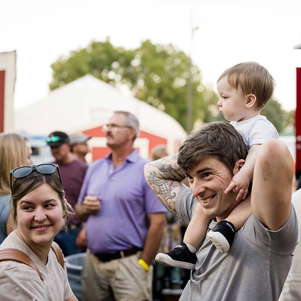 a man holds a young child on his shoulders while he and a woman smile during a crowded outdoor event at blue and gold weekend