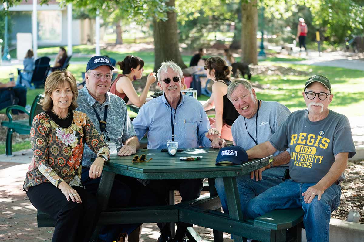 five alumni sit around a shaded picnic table outside the student center on the quad