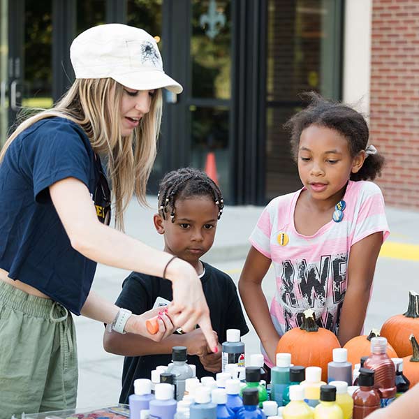 a woman helps two small children select paints for the paint your pumpkin activity outside on the student center patio