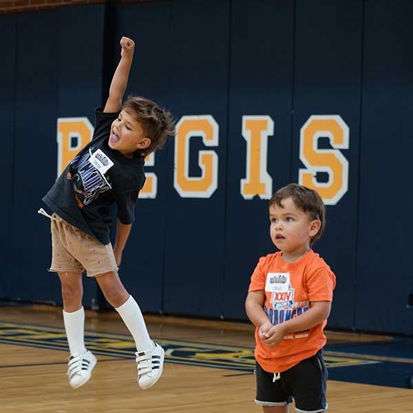 a young boy jumps with his fist in the air during an athletics event in the fieldhous. another young boy stands nearby intently watching the game.