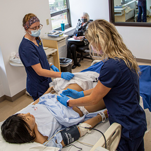 group of nursing students in a classroom performing a medical assessment on a training dummy, with the instructor observing in the background