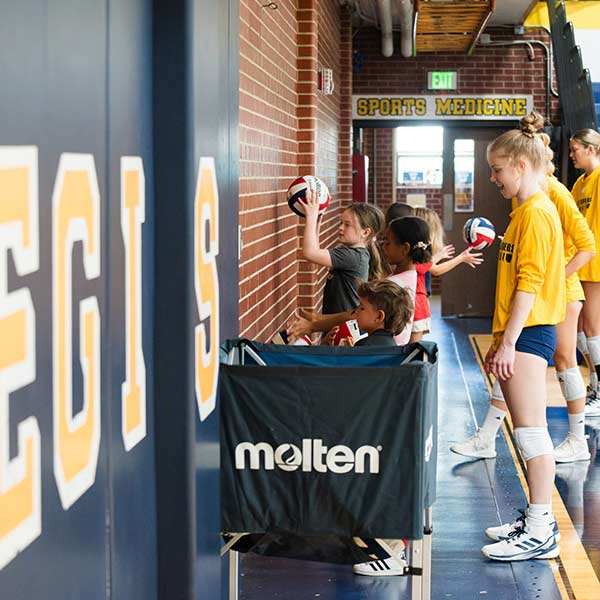 a small group of young children play with mini backetballs while Ranger athletes observe and instruct in the field house on the regis campus