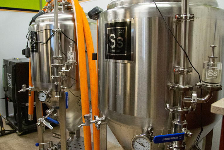 Two stainless steel Ss Brewtech fermentation tanks with gauges, valves, and temperature controls, connected by an orange hose. A black control unit sits to the left. 