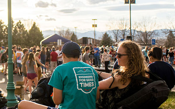 a large crowd of students enjoys an outdoor fair with tents in a parking lot on campus while students sit together on the grass