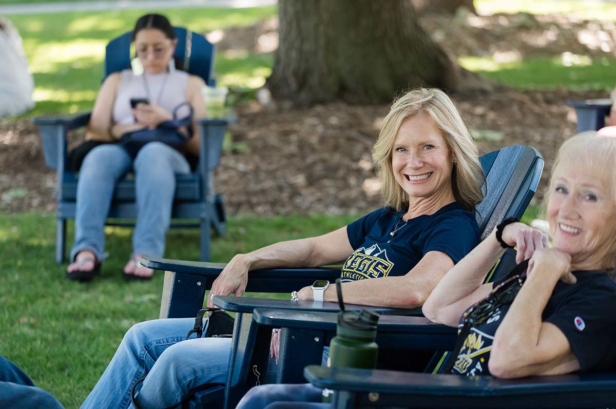 two alumni smile and lounge in blue adirondack chairs on the quad while others sit in the background