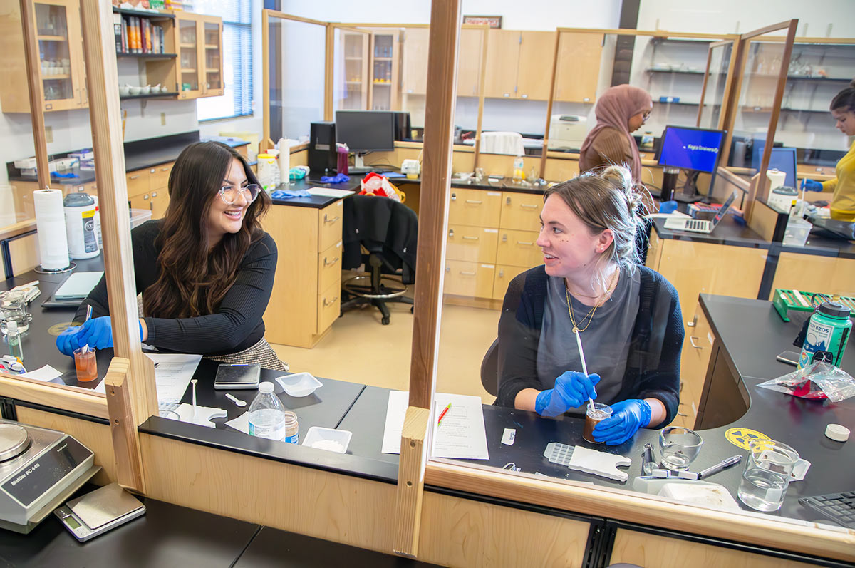 a pair of lab partners smiles at each other while working in the pharmacy lab on the regis campus