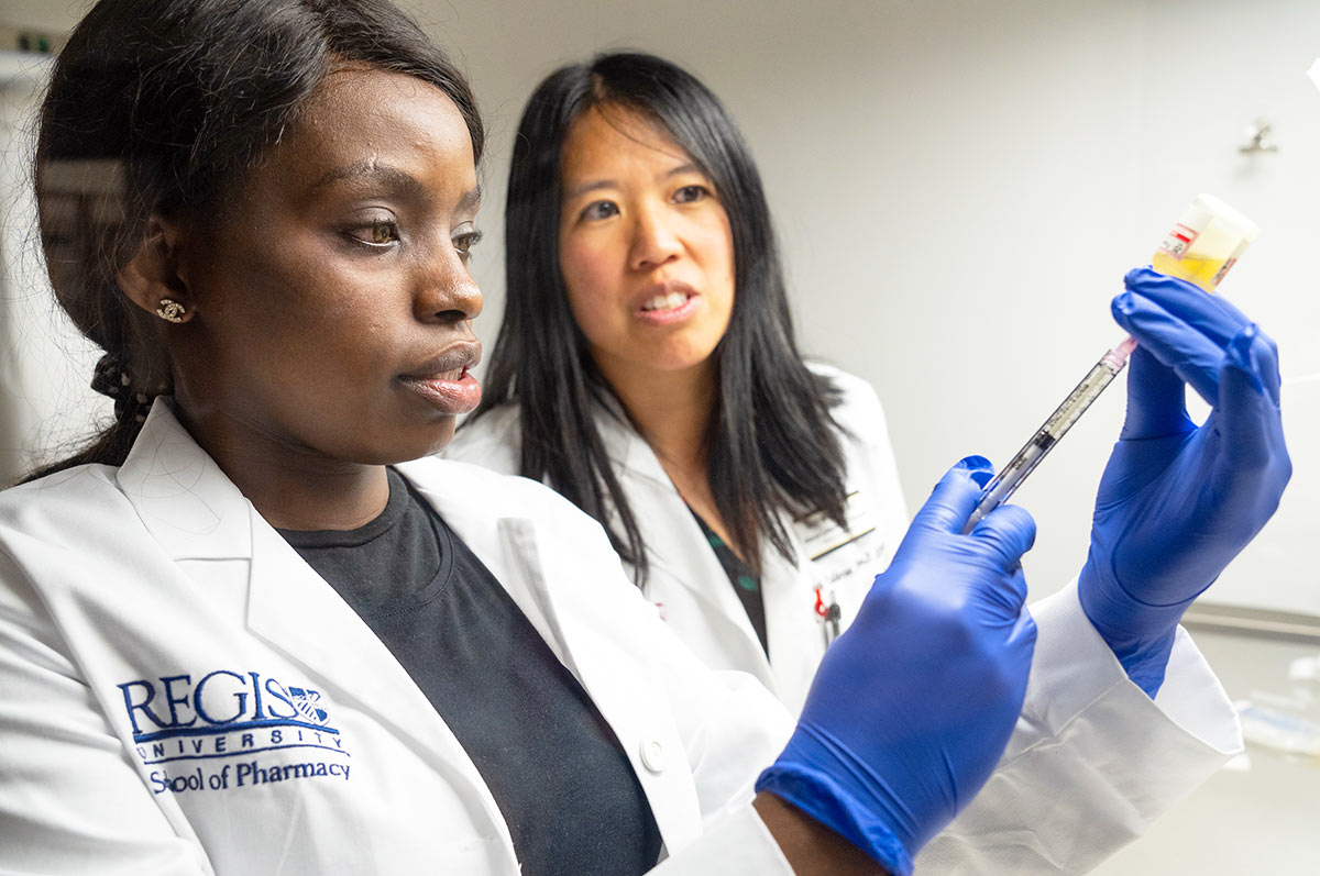 a pharmacy student draws liquid from a vial into a syringe while her professor observes in the pharmacy lab on the Regis campus