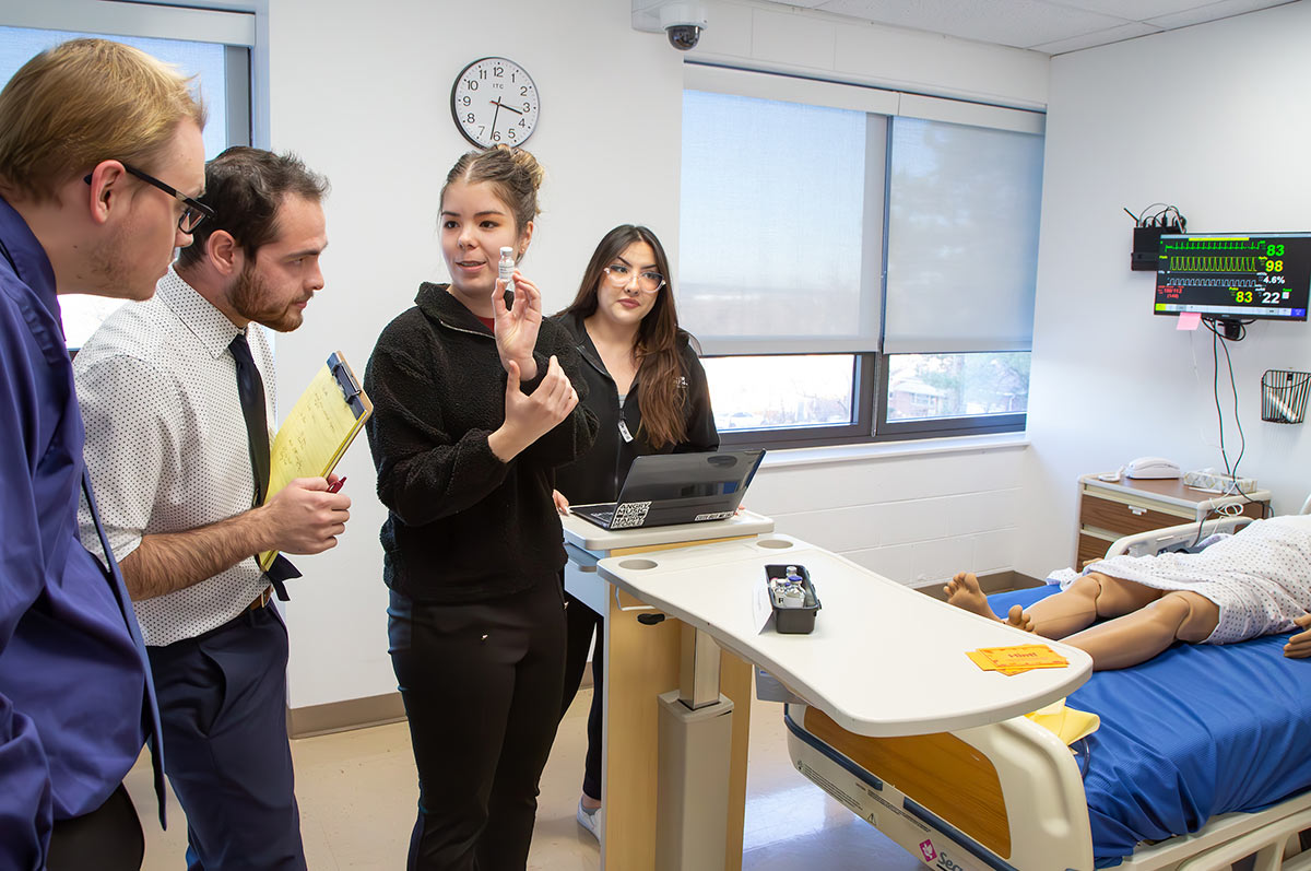 a group of students works in the simulation lab on the Regis campus. One student holds up a vial for the others to inspect while another takes notes on a laptop. A simulation dummy rests in the hospital bed nearby.