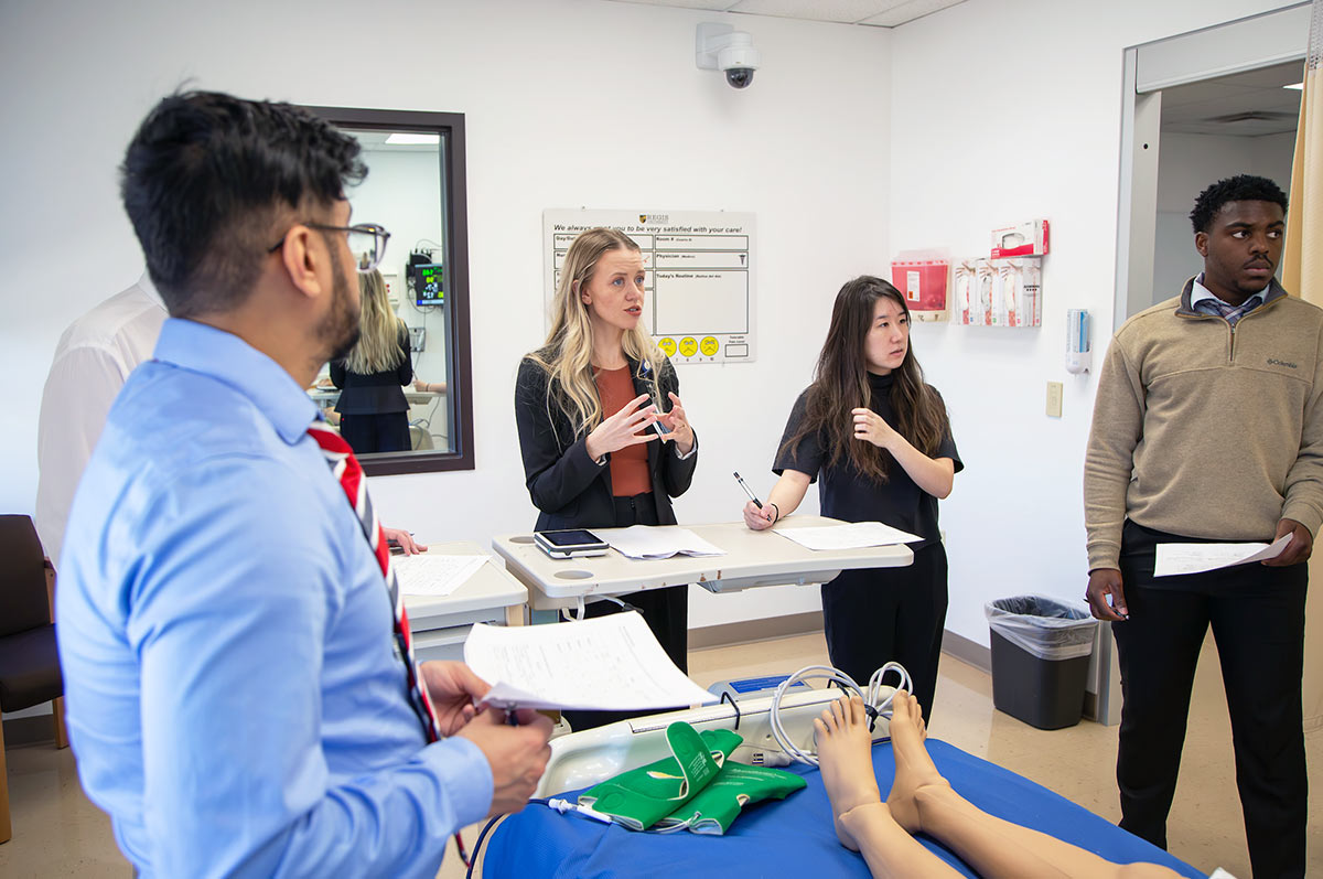 a group of pharmacy students stands around a bed holding a simulation dummy and listens intently as their professor speaks in the simulation lab on the Regis campus.