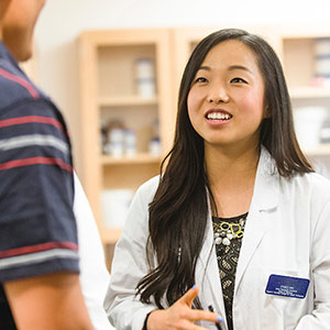 a pharmacy student in a white coat waits on a patient at the pharmacy lab on the Regis campus