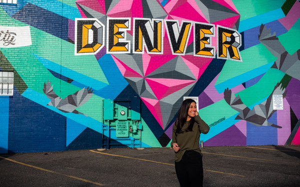 Person stands in front of the side of a building with art that reads, Denver