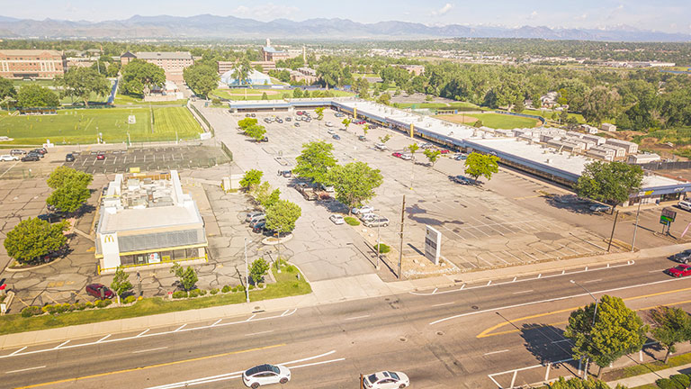 Aerial view of the current site of the proposed Regis Village development showing the now-demolished McDonalds, Regis Square parking lot and businesses with busy Federal Blvd. to the east. To the west, campus buildings are visible, including Clarke Hall and historic Main Hall. The Rocky Mountains are visible in the distant background.