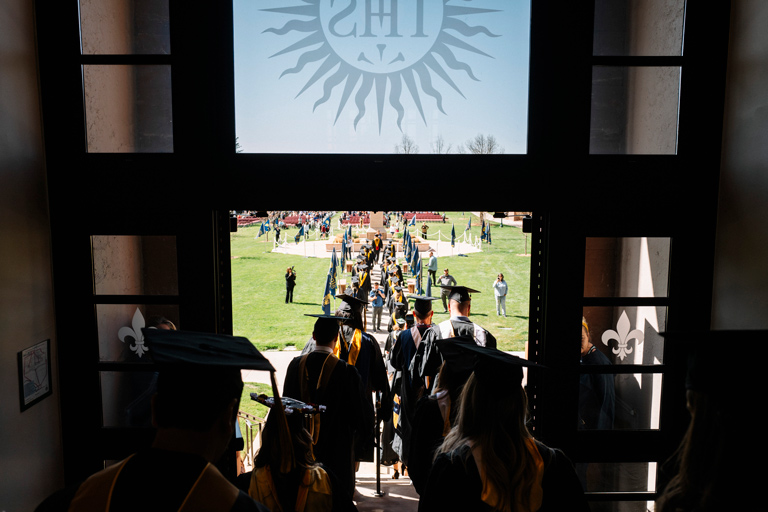 group of students dressed in cap and gown for graduation, walking down front steps of Main Hall on Denver Northwest campus