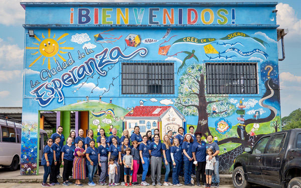 large group of students and community members posed outside of a building painted with a mural depicting children playing with kites on a grassy hill and a sign reading La Ciudad de la Esperanza