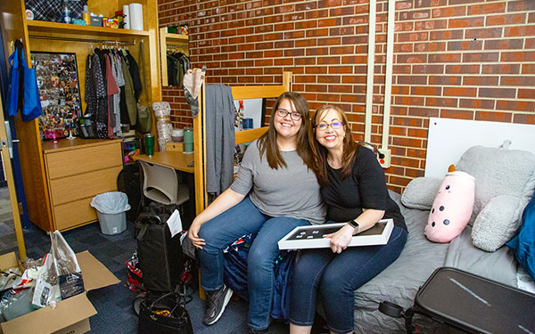 a new residential student and their parent sits on the bed in a dorm room unpacking the student's belongings